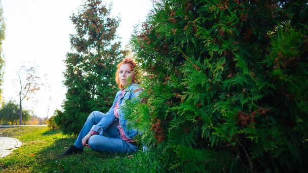Young woman with red hair among trees. Portrait of young attractive female with trendy hairstyle standing in spring garden and looking at camera - Photo, image