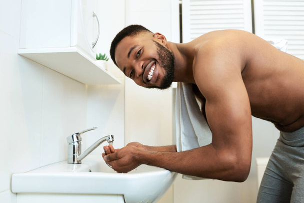 Póló nélküli afrikai Y-generációs férfi mossa az arcát a fürdőszobában - Fotó, kép