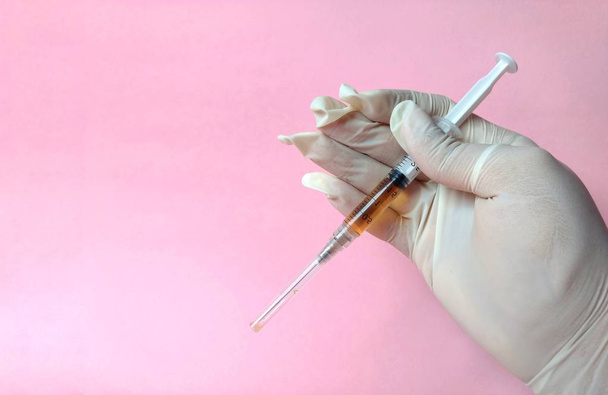 Рука с белой латексной перчаткой со шприцем готова к использованию на розовом фоне. медицинская концепция
 - Фото, изображение