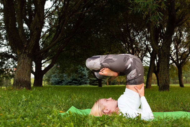 Rubia en una camiseta blanca y polainas deportivas haciendo yoga sobre una alfombra en el parque
 - Foto, imagen