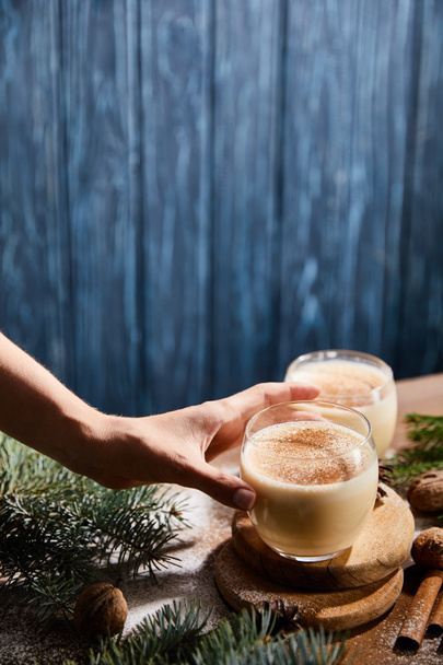 cropped view of woman taking glass with eggnog cocktail near spruce branches on blue wooden background - Foto, afbeelding