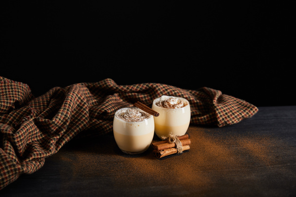 delicious eggnog cocktail, cinnamon sticks and checkered cloth on table covered with cinnamon powder isolated on black - Fotografie, Obrázek
