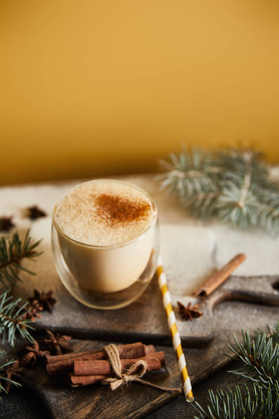 traditional eggnog cocktail on chopping board covered with sugar powder, spruce branches, cinnamon sticks and straw on orange background - Fotó, kép