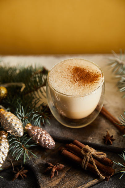 eggnog cocktail on chopping board covered with sugar powder, spruce branches, cinnamon sticks and Christmas baubles on orange background - 写真・画像