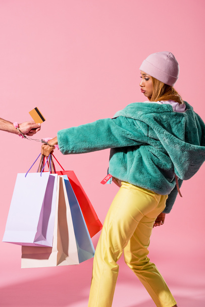 cropped view of male hand with credit card handcuffed with offended african american woman holding shopping bags on pink background, fashion doll concept - 写真・画像