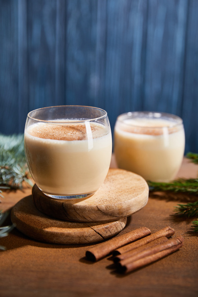 delicious eggnog cocktail on round wooden boards near spruce branches and cinnamon sticks on blue textured background  - Photo, Image