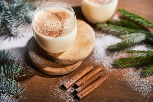 flavored eggnog cocktail on round boards near spruce branches and cinnamon sticks on wooden table covered with sugar powder - Zdjęcie, obraz