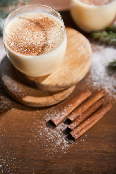 flavored eggnog cocktail on round boards near spruce branches and cinnamon sticks on wooden table covered with sugar powder - Photo, Image