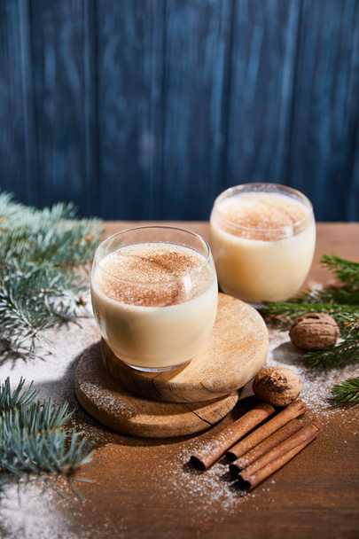delicious eggnog cocktail, spruce branches, cinnamon sticks and walnuts on wooden table covered with sugar powder on blue textured background - Photo, Image