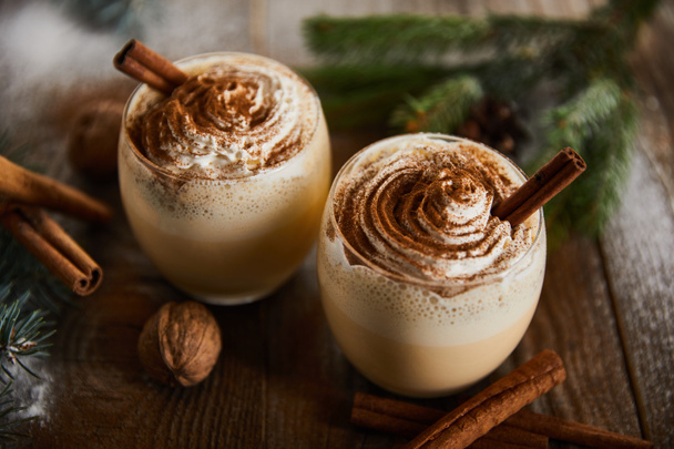 traditional eggnog cocktail with whipped cream near cinnamon sticks, spruce branches and walnuts on wooden table - Foto, afbeelding