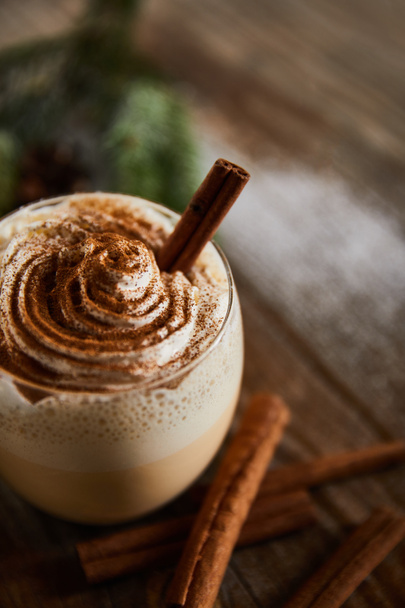 close up view of flavored eggnog drink with whipped cream on wooden table near cinnamon sticks - Foto, imagen