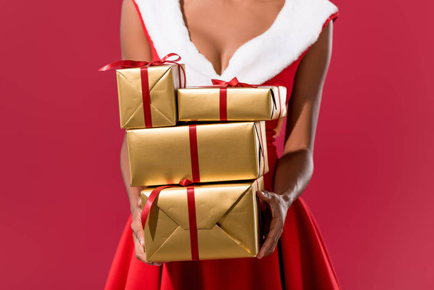 cropped view of african american girl in santa hat and christmas dress holding gift boxes isolated on red - 写真・画像