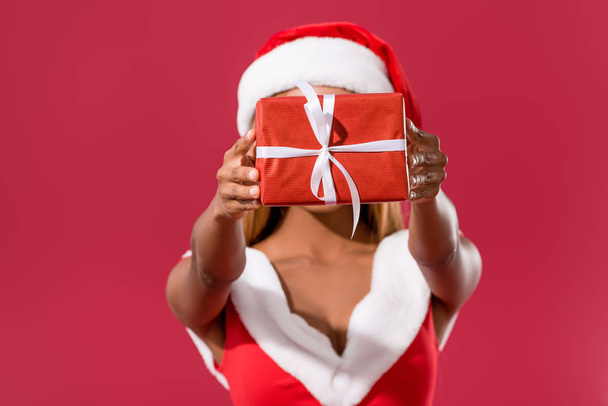 giovane ragazza afroamericana in cappello di Babbo Natale e vestito di Natale in possesso di scatola regalo di fronte al viso isolato sul rosso
 - Foto, immagini