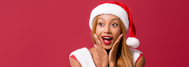 panoramic shot of african american girl in santa hat holding hands near face isolated on red - Фото, изображение