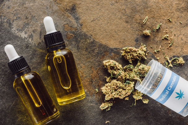 vrchní pohled na lékařské marihuanové pupeny v blízkosti kontejneru a láhve s konopným olejem na mramorovém povrchu - Fotografie, Obrázek