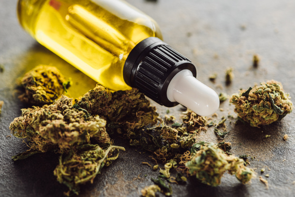 στενή άποψη της ιατρικής μαριχουάνας μπουμπούκια κοντά μπουκάλι με λάδι κάνναβης στην επιφάνεια του μαρμάρου - Φωτογραφία, εικόνα