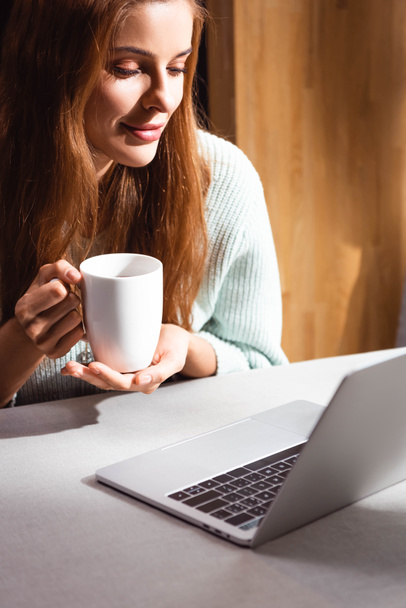 hymyilevä punapää nainen kupin kahvia kannettavan tietokoneen avulla kahvilassa
 - Valokuva, kuva