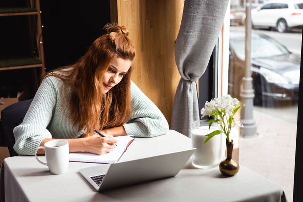 rothaarige Frau schreibt in Notizblock, während sie Webinar auf Laptop im Café anschaut - Foto, Bild