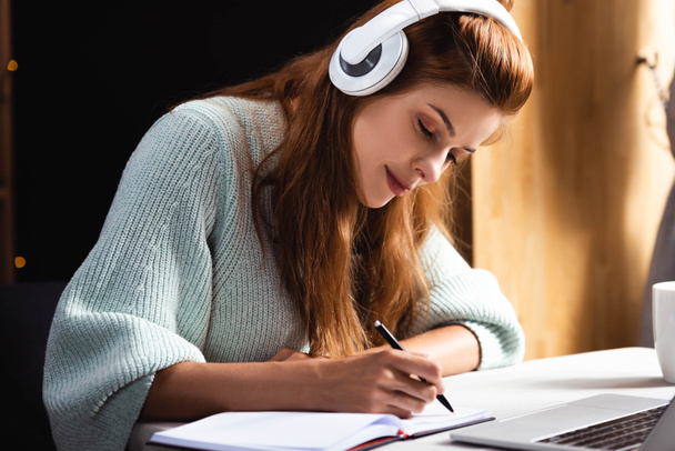 γυναίκα στα ακουστικά γράφοντας και μελετώντας σε απευθείας σύνδεση με το lap-top στο καφέ - Φωτογραφία, εικόνα