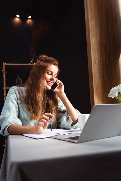 mujer sonriente hablando en el teléfono inteligente mientras estudia en línea con el ordenador portátil en la cafetería
 - Foto, imagen