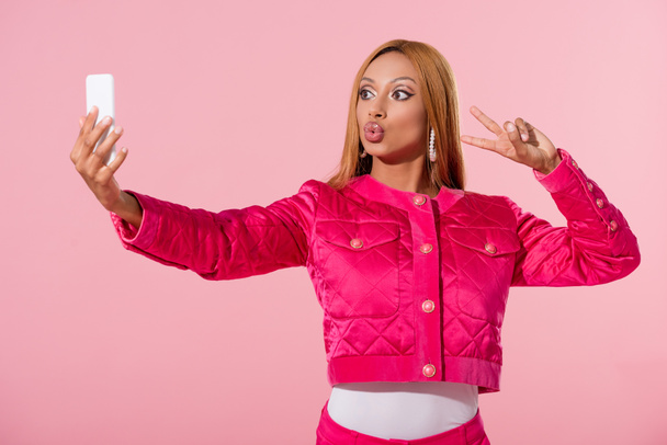 stylish african american woman showing victory gesture and taking selfie isolated on pink, fashion doll concept - Photo, Image