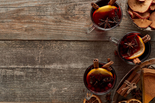 bovenaanzicht van stuk taart en rode gekruide glühwein met bessen, anijs, sinaasappelschijfjes en kaneel op houten rustieke tafel - Foto, afbeelding