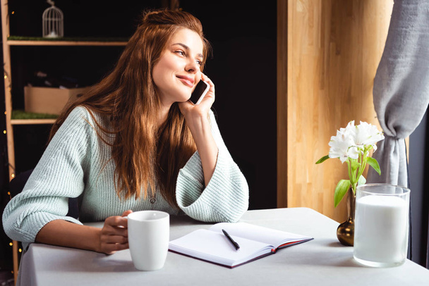 uśmiechnięta kobieta rozmawiająca na smartfonie w kawiarni z notatnikiem i filiżanką kawy - Zdjęcie, obraz