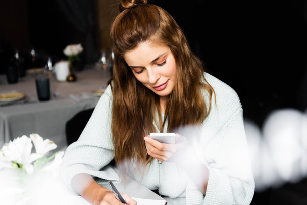 beautiful woman using smartphone and writing in notepad in cafe with cup of coffee - 写真・画像