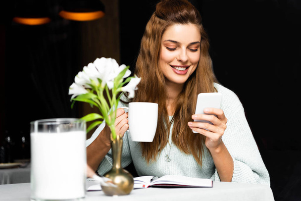 smiling woman with cup of coffee using smartphone in cafe with notepad  - Zdjęcie, obraz