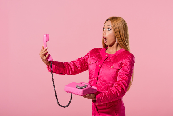 zszokowana, modna afrykańska kobieta patrząca na telefon odizolowany od różowej, modnej koncepcji lalki - Zdjęcie, obraz