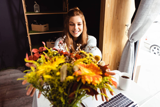 улыбающаяся рыжая женщина с букетом осенних цветов в кафе с ноутбуком и чашкой кофе
  - Фото, изображение