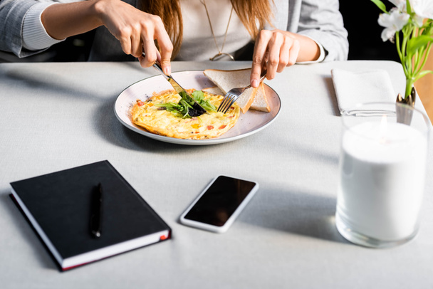cropped view of woman eating omelet for breakfast in cafe with notepad and smartphone - Foto, Imagem