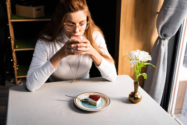 attractive girl drinking coffee with cake in cafe with flowers in vase - Фото, изображение