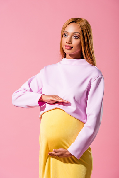 trendy, zwangere Afrikaans-Amerikaanse vrouw die de buik aanraakt en geïsoleerd kijkt op roze, modepoppenconcept - Foto, afbeelding