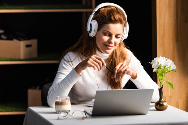 κοκκινομάλλα χαμογελαστή γυναίκα με ακουστικά βλέποντας webinar στο φορητό υπολογιστή στο καφέ - Φωτογραφία, εικόνα