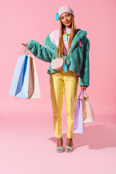 sonriente, elegante mujer afroamericana sosteniendo bolsas de compras sobre fondo rosa, concepto de muñeca de moda
 - Foto, imagen