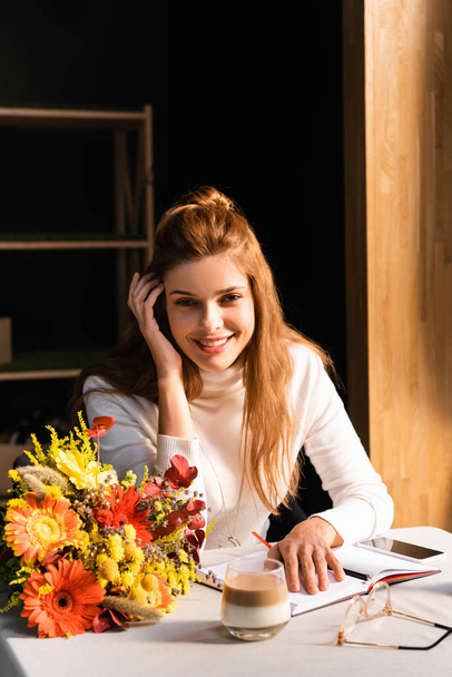 attractive woman with bouquet of autumn flowers in cafe with smartphone, cup of coffee and notebook - Zdjęcie, obraz
