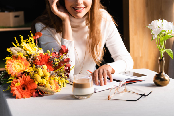 cropped view of woman with bouquet of autumn flowers in cafe with smartphone and notebook - Fotografie, Obrázek
