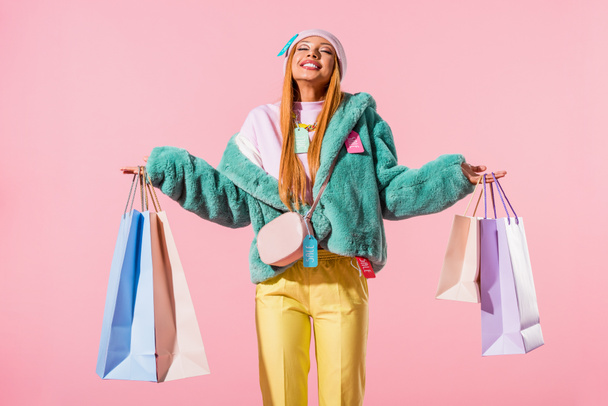 幸せなスタイリッシュなアフリカ系アメリカ人女性が目を閉じてピンクのファッションドールのコンセプトで隔離されたショッピングバッグを持って立って - 写真・画像