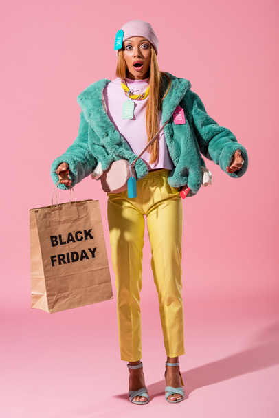 schockiertes, stylisches afrikanisches ameikanisches Mädchen mit Einkaufstasche mit schwarzer Freitag-Aufschrift auf rosa Hintergrund, Modepuppenkonzept - Foto, Bild