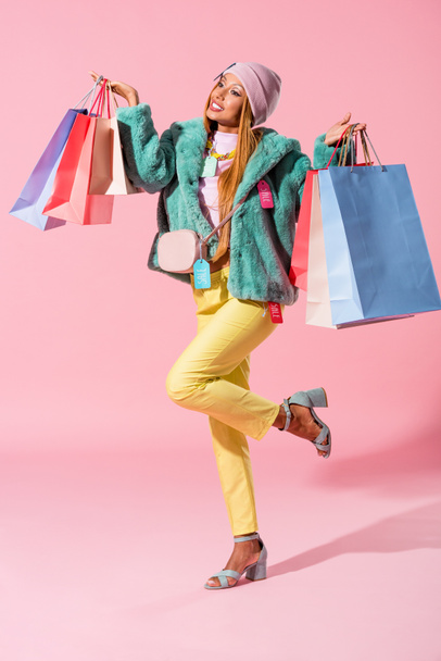 cheerful, stylish african american woman holding shopping bags while standing on one leg on pink background, fashion doll concept - Фото, зображення