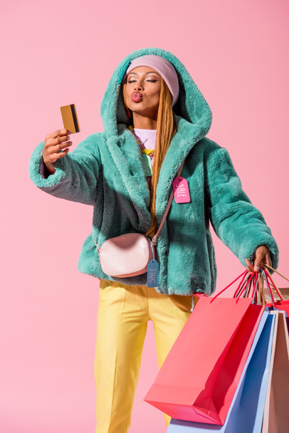 μοντέρνα αφροαμερικάνικη γυναίκα με πρόσωπο πάπιας που κρατάει πιστωτική κάρτα και τσάντες για ψώνια απομονωμένες σε ροζ, fashion doll concept - Φωτογραφία, εικόνα
