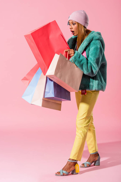 překvapený, módní africký Američan žena při pohledu v nákupní tašce na růžovém pozadí, módní panenka koncept - Fotografie, Obrázek