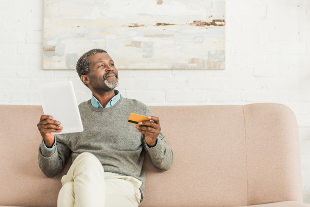 senior african american man holding digital tablet and credit card while sitting on sofa and looking away - Photo, Image