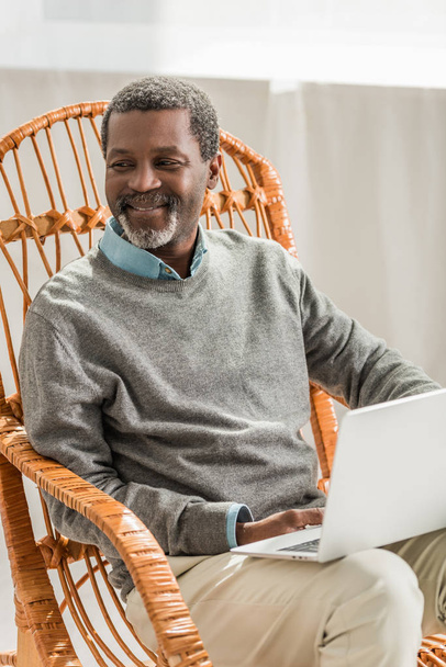smiling african american man sitting in wicker chair with laptop  - Фото, зображення