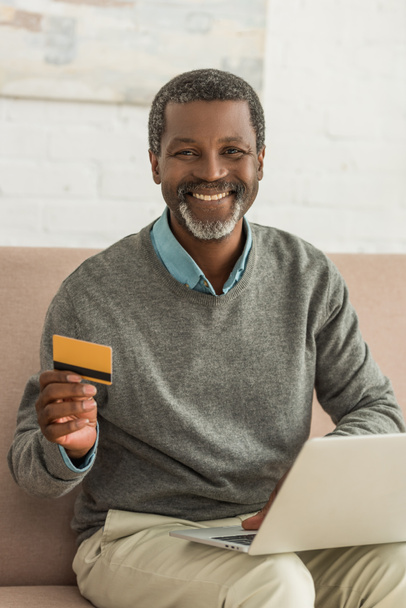 senior african american man sitting on sofa with laptop, holding credit card and smiling at camera - Foto, Bild