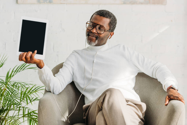 hombre afroamericano elegante sentado en sillón, escuchando música en auriculares y sosteniendo la tableta digital
 - Foto, imagen