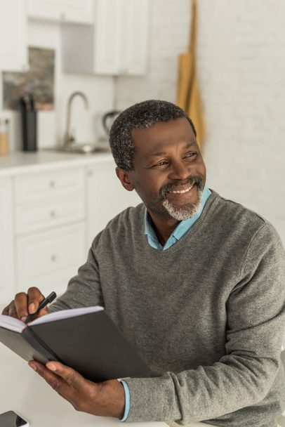 hombre afroamericano mayor sonriendo y mirando hacia otro lado mientras escribe en un cuaderno
 - Foto, Imagen