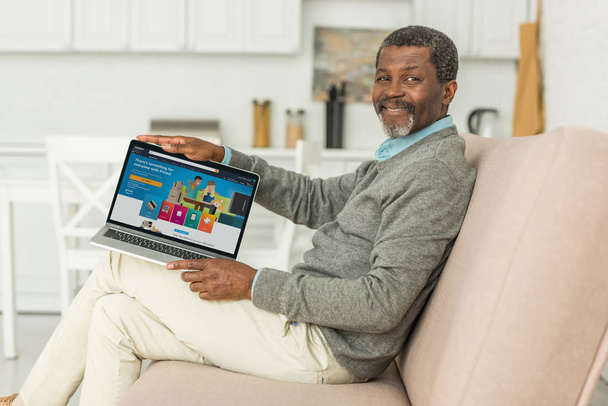 KYIV, UKRAINE - OCTOBER 2, 2019: Cheerful african american man sitting on sofa and showing laptop with Amazon app on screen. - Photo, Image