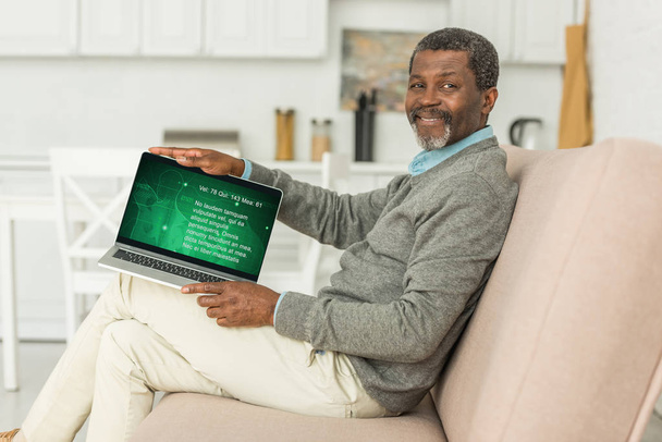 KYIV, UCRANIA - 2 DE OCTUBRE DE 2019: Hombre afroamericano alegre sentado en el sofá y mostrando el ordenador portátil con información médica en la pantalla
. - Foto, imagen